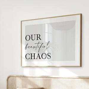May include: A framed wall art print featuring the text OUR beautiful CHAOS in black lettering against a light grey background. The print is displayed above a bed with a cane headboard, green bedding, and a tan throw blanket in a bright room.