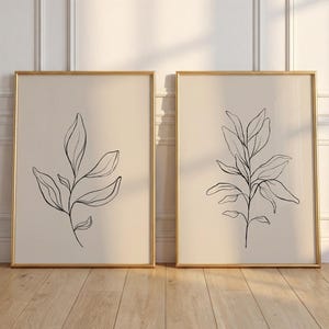 Op de afbeelding: Twee rechthoekige wandkunstprints in dunne gouden lijsten. Elke print heeft een beige achtergrond met een zwarte lijntekening van een bebladerde plantenstengel. De kunstwerken staan naast elkaar en tonen botanische illustraties met strakke lijnen.