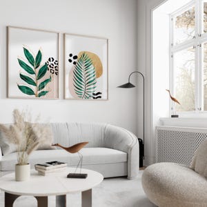 May include: Two framed botanical art prints with gold accents and abstract shapes hang above a white sofa. The prints feature green leaves, leopard print, and gold and black shapes. A modern living room setting.