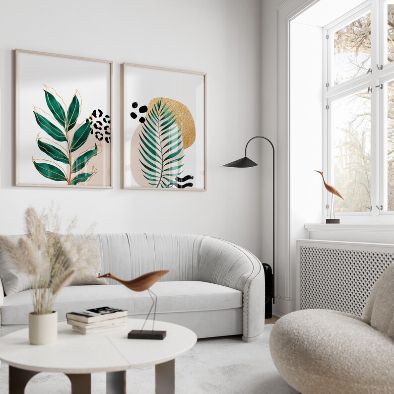 May include: Two framed botanical art prints with gold accents and abstract shapes hang above a white sofa. The prints feature green leaves, leopard print, and gold and black shapes. A modern living room setting.