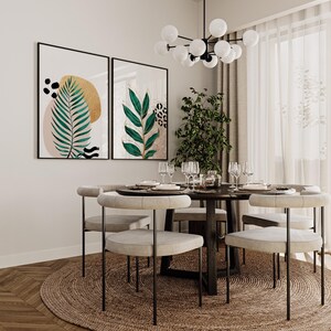 May include: A dining room scene featuring a round wooden table set for a meal, surrounded by light-colored chairs with black metal legs. Two framed botanical art prints hang on the wall. A jute rug sits beneath the table, and a modern chandelier hangs above.