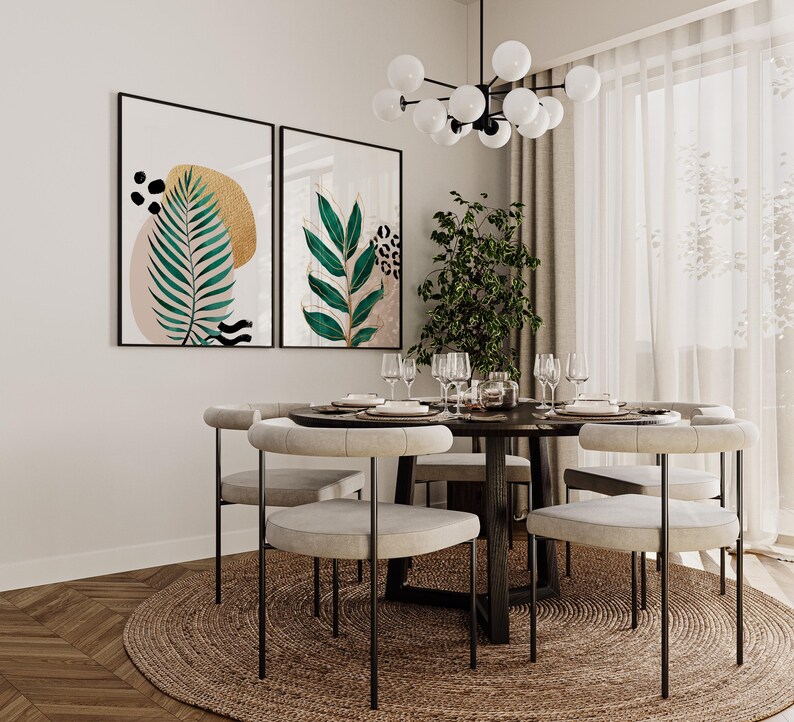 May include: A dining room scene featuring a round wooden table set for a meal, surrounded by light-colored chairs with black metal legs. Two framed botanical art prints hang on the wall. A jute rug sits beneath the table, and a modern chandelier hangs above.