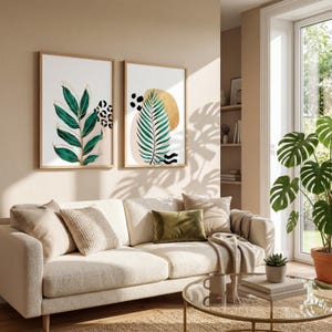 May include: Two framed botanical art prints. One features green leaves with gold accents and a leopard print. The other showcases a green palm leaf, gold circle, and black abstract shapes. The prints hang above a cream-colored sofa.