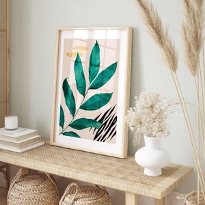 a framed artwork of a green leaf with black stripes, placed on a wooden table. The table also holds a white vase with white flowers and a stack of books.
