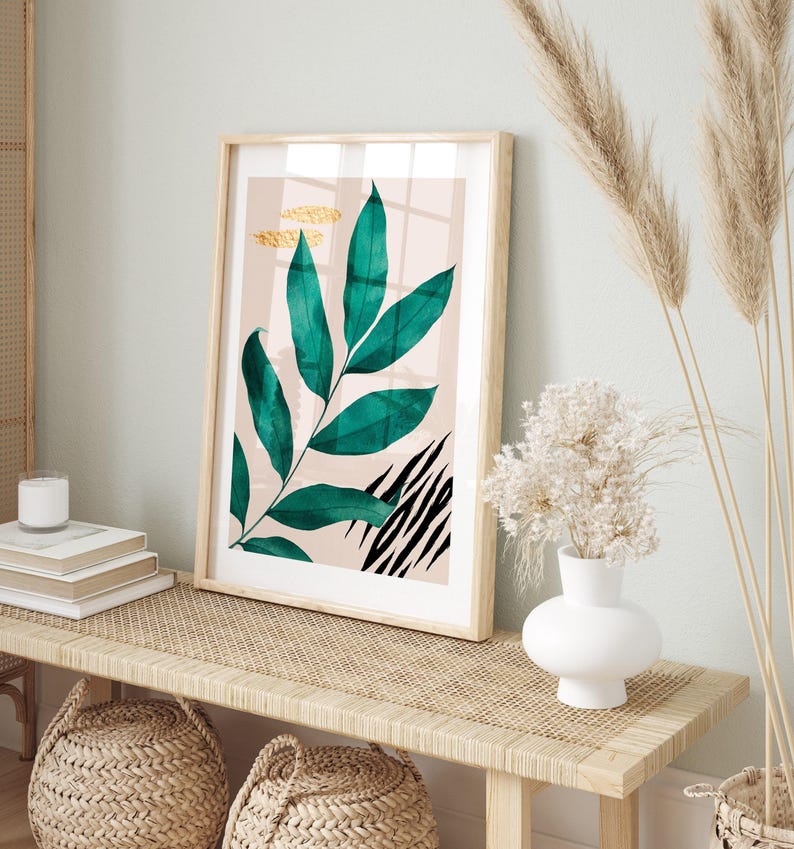 a framed artwork of a green leaf with black stripes, placed on a wooden table. The table also holds a white vase with white flowers and a stack of books.