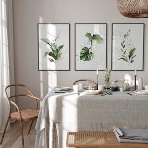 a dining room with a table set for a meal, featuring a tablecloth, plates, cups, and a vase with flowers. Three framed prints of green plants are displayed on the wall above the table.