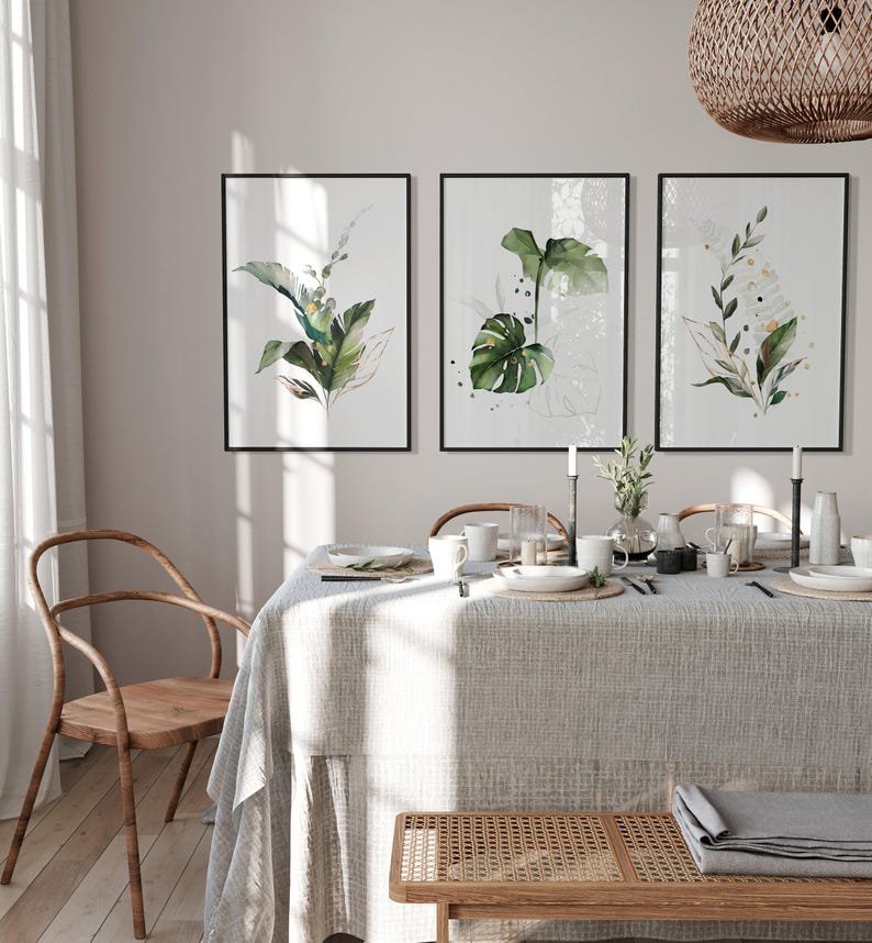 a dining room with a table set for a meal, featuring a tablecloth, plates, cups, and a vase with flowers. Three framed prints of green plants are displayed on the wall above the table.