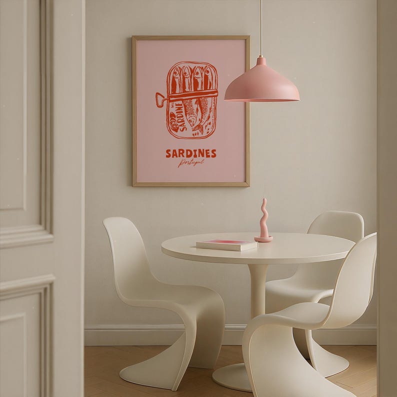 a dining area with a white table and chairs, a pink lamp hanging above the table, and a framed poster of a can of Sardines on the wall.