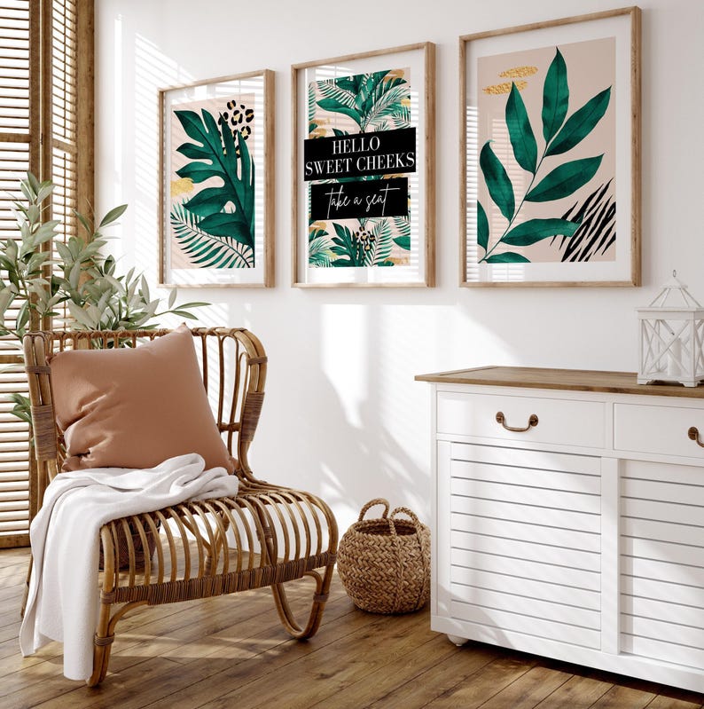 a cozy living room with a wicker chair, a woven basket, and three framed prints on the wall. The prints include tropical leaves and the text "Hello Sweet Cheeks Take a seat".