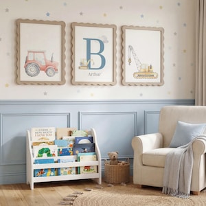 May include: Three framed wall art prints featuring a red tractor, a large blue letter B with a small excavator, and a yellow crane. Below the prints is a white wooden bookshelf filled with children's books, a wicker basket, and an armchair.