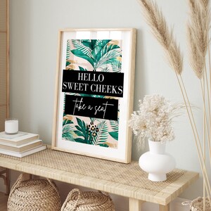 a framed poster with a tropical leaf pattern and the text "Hello Sweet Cheeks Take a Seat" on a wooden table.