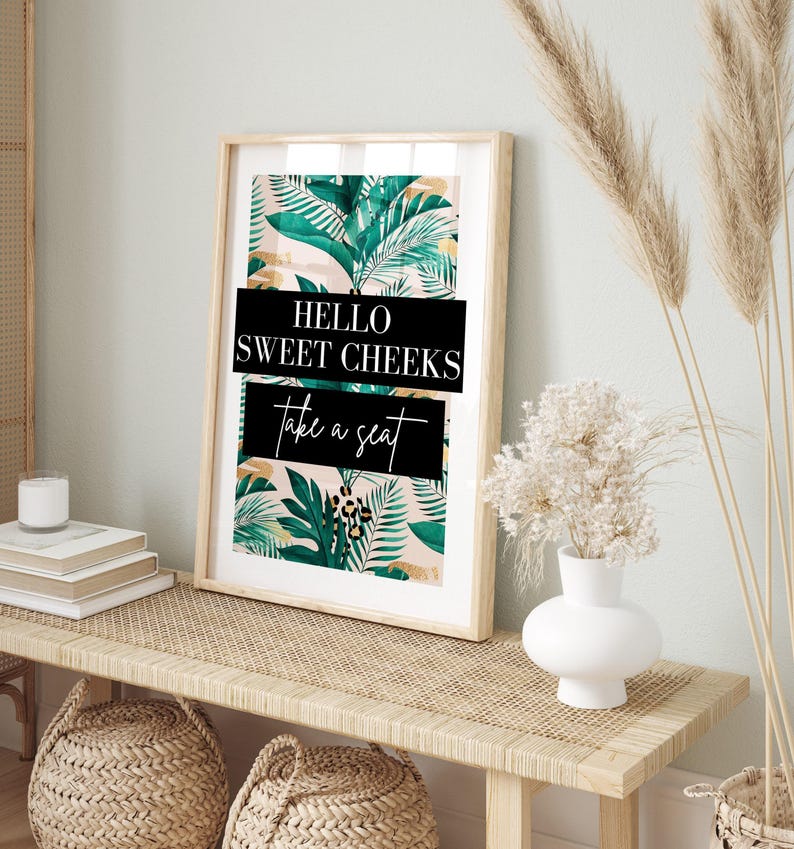 a framed poster with a tropical leaf pattern and the text "Hello Sweet Cheeks Take a Seat" on a wooden table.