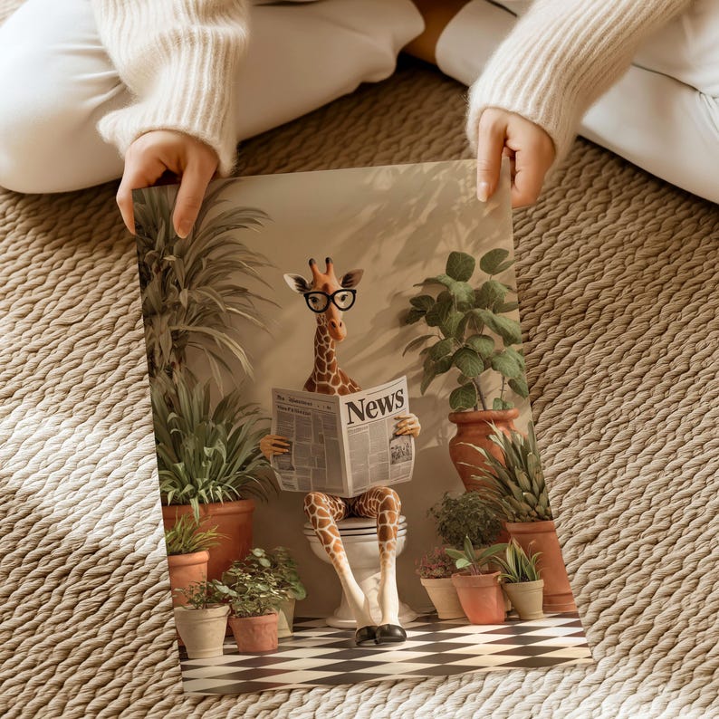 A person is holding a framed print featuring a giraffe sitting on a toilet and reading a newspaper, surrounded by potted plants.