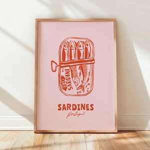 a framed poster with a pink background and a red illustration of a can of sardines. The poster has the text "SARDINES" and "Portugal" written on it.