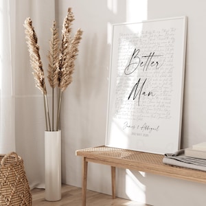 a white framed poster with the text "Better Man" and "James & Affigail" written on it, placed on a wooden bench in a room with a woven basket and a tall vase with dried reeds.
