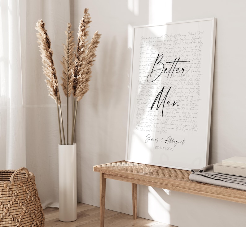 a white framed poster with the text "Better Man" and "James & Affigail" written on it, placed on a wooden bench in a room with a woven basket and a tall vase with dried reeds.