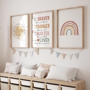 May include: Three framed wall art prints featuring a sun, a rainbow, and the text: You are BRAVER Than you believe STRONGER Than you seem SMARTER Than you think and LOVED More than you know. A string of fabric bunting hangs below the frames.