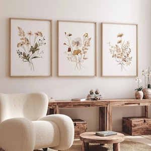 Puede incluir: Tres láminas artísticas botánicas enmarcadas con ilustraciones en acuarela de flores secas en tonos neutros. Las láminas se exhiben en una pared blanca sobre una consola de madera, creando una estética serena y natural.