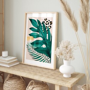 a framed artwork of a green leaf with a leopard print design, displayed on a wooden table. The artwork is accompanied by a white vase with white flowers, a stack of books, and a woven basket.