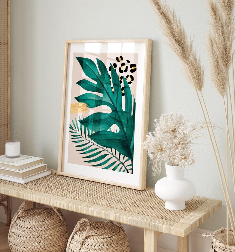 a framed artwork of a green leaf with a leopard print design, displayed on a wooden table. The artwork is accompanied by a white vase with white flowers, a stack of books, and a woven basket.