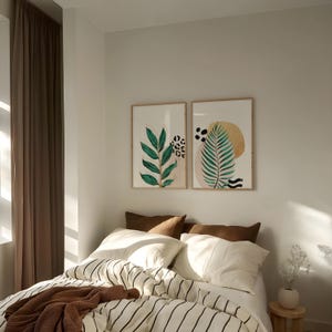 May include: Two framed botanical art prints with green leaves and abstract shapes hang above a bed. The bed has white bedding with black stripes, brown pillows, and a brown throw blanket. A small wooden stool holds a vase with white flowers.