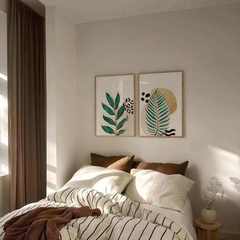 May include: Two framed botanical art prints with green leaves and abstract shapes hang above a bed. The bed has white bedding with black stripes, brown pillows, and a brown throw blanket. A small wooden stool holds a vase with white flowers.