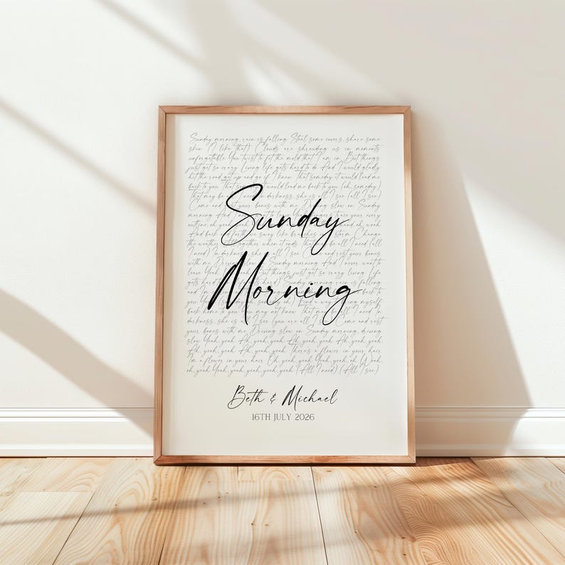 A framed print with the text "Sunday Morning" and the names "Beth & Michael" printed on it, placed on a wooden floor against a white wall.