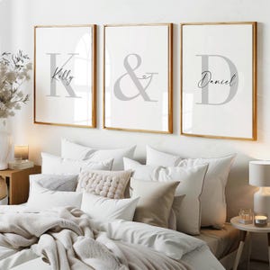Pode incluir: Três quadros de arte de parede com molduras douradas. A impressão da esquerda apresenta uma letra K grande em cinzento com o nome Kelly. A impressão central mostra um grande e comercial cinzento com um pequeno coração. A impressão da direita mostra uma letra D grande em cinzento com o nome Daniel.
