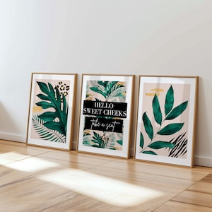 Three framed prints with tropical plant designs are displayed on a wooden floor against a white wall.