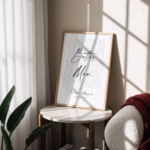 a framed poster with the text "Better Man" on it, placed on a round table next to a plant and a stack of magazines.