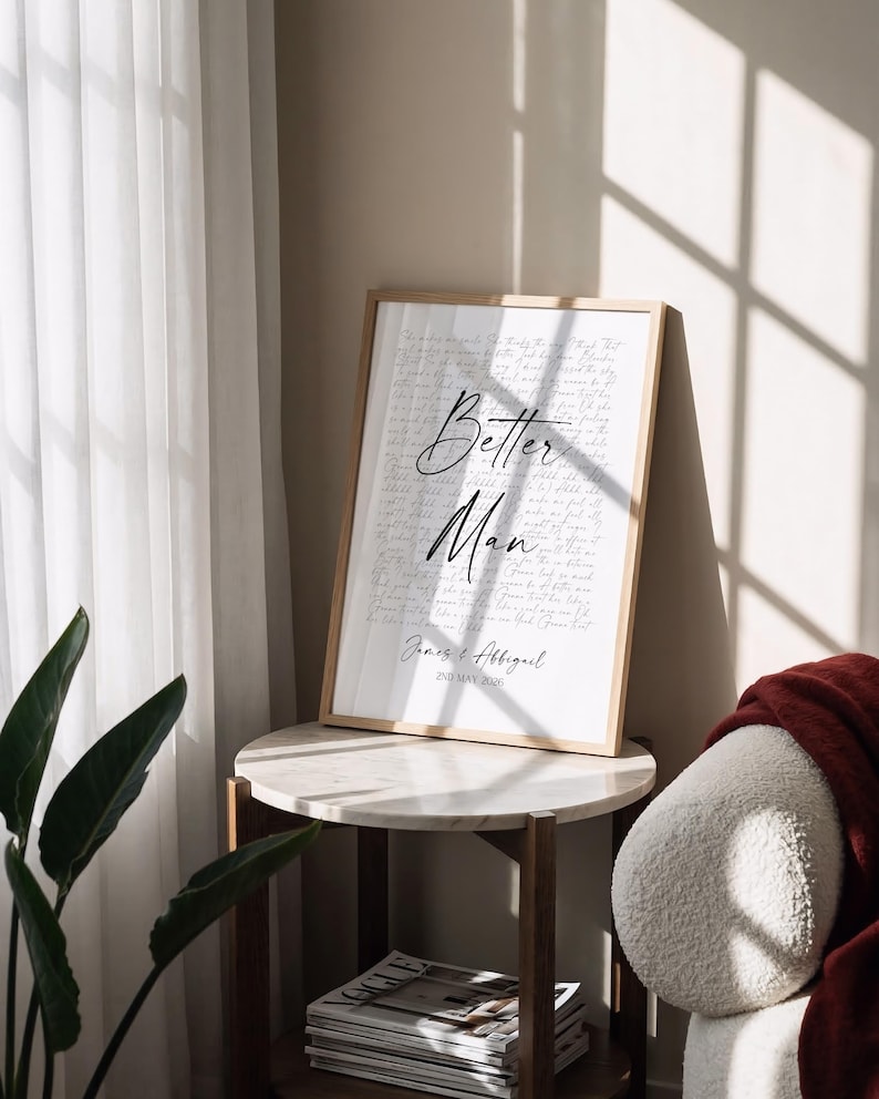 a framed poster with the text "Better Man" on it, placed on a round table next to a plant and a stack of magazines.