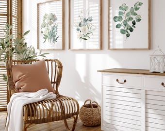 May include: Three framed botanical prints with watercolor floral designs hang on a white wall. The prints feature flowers and greenery in shades of green, white, and blue. A wicker chair with a brown pillow and white blanket sits in front of a window.