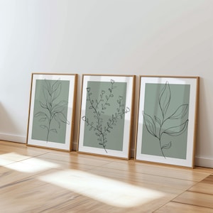 May include: Three framed botanical art prints. Each print features a different black line drawing of leaves and stems on a sage green background. The frames are a light brown colour, and the prints are displayed on a wooden floor.