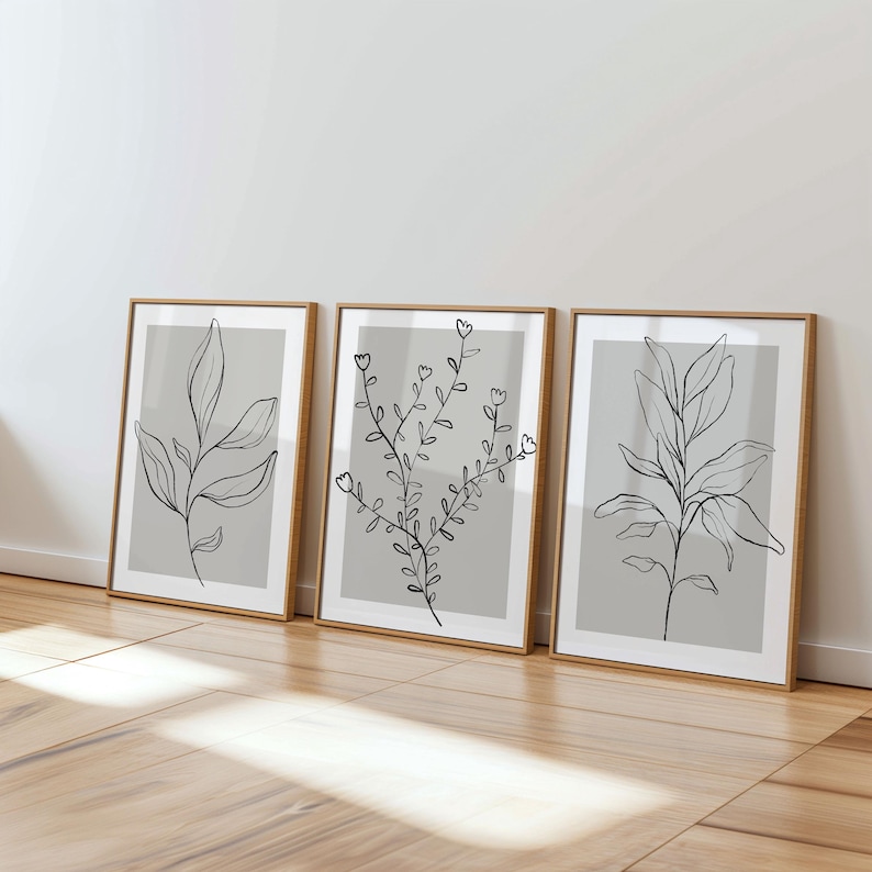 Three framed artworks depicting abstract plant designs are displayed on a wooden floor against a white wall.