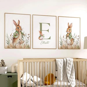 Pode incluir: Três impressões em aquarela emolduradas com ilustrações de Peter Rabbit. As impressões retratam coelhos em um campo, um com a letra "E" e o nome "Elliott". A obra de arte tem uma paleta de cores suaves de verdes, marrons e bege, ideal para um berçário.