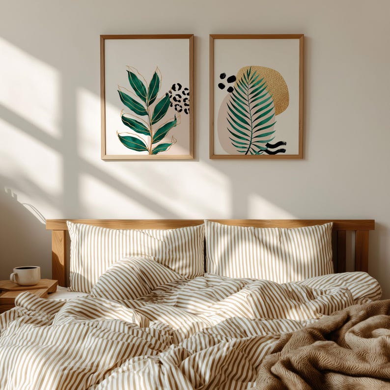 May include: Two framed botanical art prints with green and gold leaf designs hang above a bed with striped bedding. The bed has a wooden headboard and a brown blanket. A small wooden tray with a mug is on the nightstand.