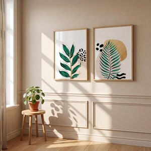 May include: Two framed botanical art prints. One features a green leaf design with gold accents and a leopard print pattern. The other print has a green palm leaf, gold circle, and abstract black shapes. Both are in light wood frames.