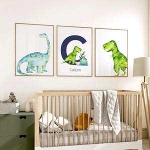 Könnte beinhalten: Drei gerahmte Aquarelldrucke mit Dinosauriern. Die Drucke zeigen einen blau-grünen Brontosaurus, einen grünen Dinosaurier mit dem Buchstaben "C" und dem Namen "Callum" sowie einen grünen T-Rex. Die Drucke werden über einem Kinderbett ausgestellt.