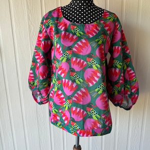 May include: A green and pink blouse with a colourful bird and flower pattern. The blouse has a round neckline and 3/4 length sleeves.