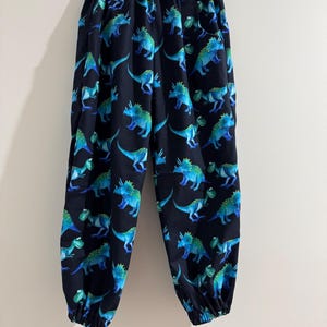 May include: Black trousers featuring a repeating pattern of blue and green dinosaurs. The trousers have an elasticated waistband and cuffs. The dinosaurs are depicted in different poses on a dark background.