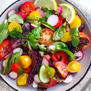 May include: A colourful salad with red and yellow tomatoes, cucumbers, radishes, and leafy greens in a white bowl with a black rim.