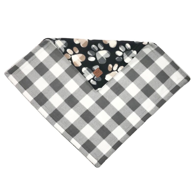 Puede incluir: Una manta reversible para perros con un patr&oacute;n de cuadros de b&uacute;falo blanco y negro en un lado y un vell&oacute;n negro con huellas de patas y corazones en el otro lado.
