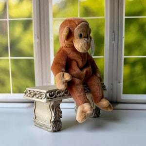 May include: A plush monkey toy with light brown fur on its face, hands, and feet, and darker brown fur on its body. The stuffed animal is sitting in a profile position, featuring a small tag attached to its ear and a soft, rounded appearance.