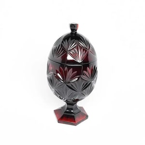 May include: A ruby red, lidded candy dish with a geometric pattern. The dish is egg-shaped and sits on a hexagonal base. The lid has a small, rounded knob. The dish is made of glass and has a vintage aesthetic.