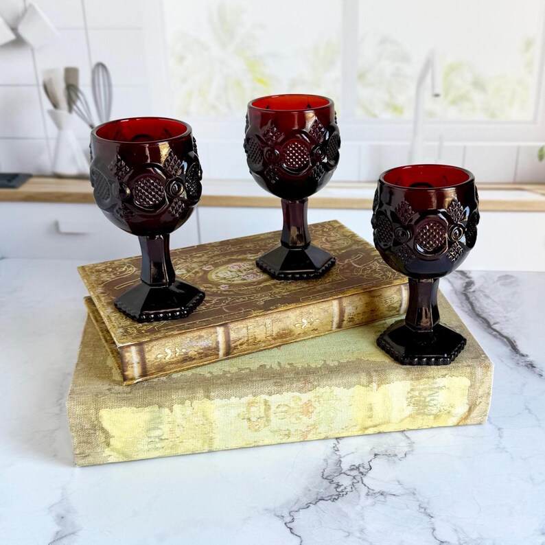 May include: Three ruby red vintage wine glasses with ornate floral and geometric designs. The glasses are displayed on top of two antique books with aged covers. The glasses have a hexagonal base and a detailed pattern around the bowl.