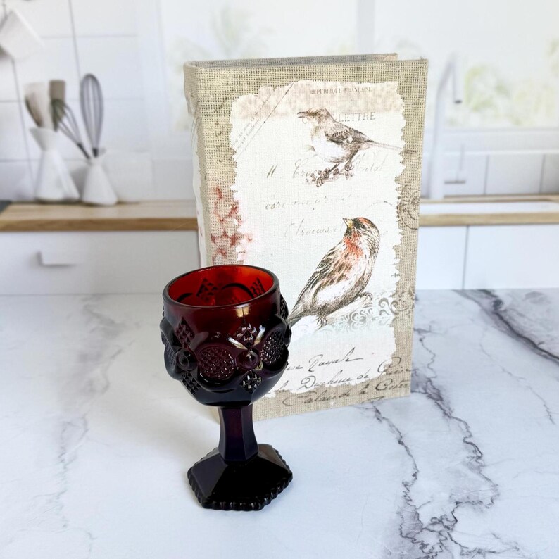 May include: A ruby red vintage wine glass with a detailed grape design. The glass sits in front of a decorative book with bird illustrations and French script. The glass has a hexagonal base.