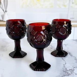 May include: Three ruby red vintage wine glasses. Each glass features a detailed embossed pattern around the bowl and a faceted base. The glasses are arranged on a marble surface, showcasing their rich color and design.