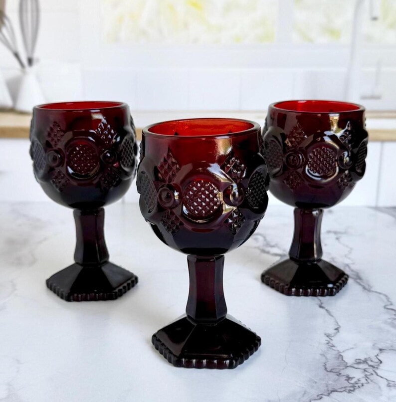 May include: Three ruby red vintage wine glasses. Each glass features a detailed embossed pattern around the bowl and a faceted base. The glasses are arranged on a marble surface, showcasing their rich color and design.