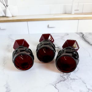 May include: Three ruby red vintage goblets with a textured design. The goblets have a faceted top and a detailed pattern around the bowl. The stemware is arranged on a white marble surface.
