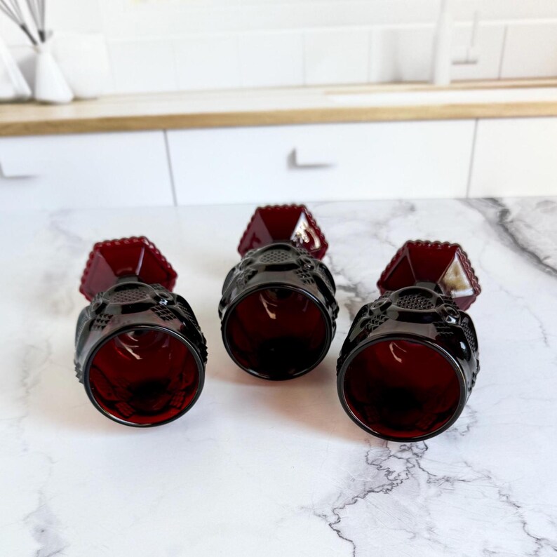 May include: Three ruby red vintage goblets with a textured design. The goblets have a faceted top and a detailed pattern around the bowl. The stemware is arranged on a white marble surface.